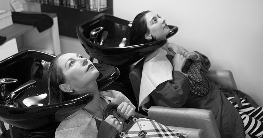 A woman and her daughter sitting at a hair washing station in a salon, gripping their purses against their chests.