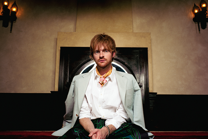 Finneas seated facing the camera, wearing a white dress shirt, scarf tied around the neck and a coat draped over his shoulders.
