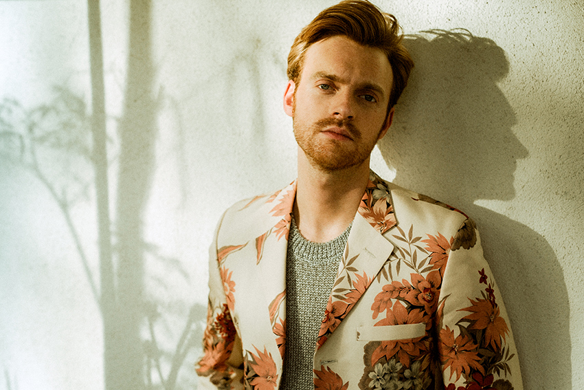 Finneas standing against a wall in a earth-toned floral blazer.