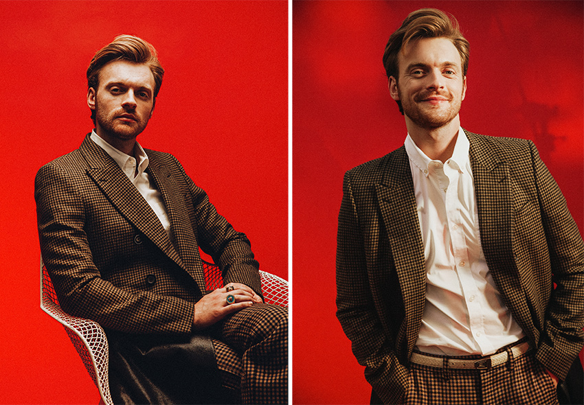 Two photos of Finneas in a suit, one sitting in a chair with his hands clasped over one thigh, the other standing with his hands in his pants pockets.