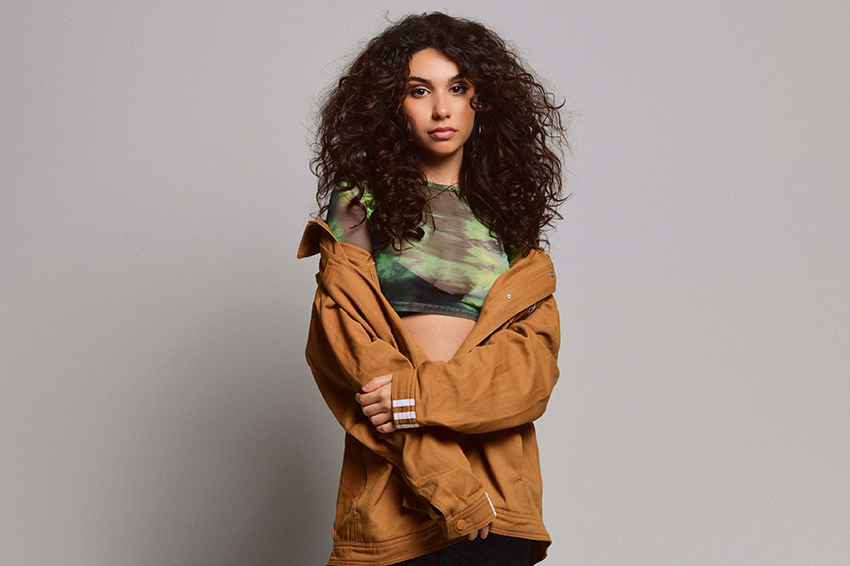 Alessia Cara posing against a grey backgroup with a camel brown coat slouched just off her shoulders.
