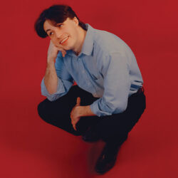 Peter McPoland crouched down with his cheek resting on his right fist, against a red backdrop.