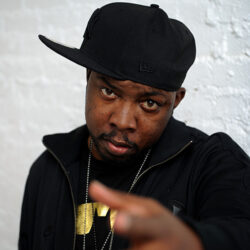 Phife facing the camera, dressed all in black with a black baseball cap worn sideways, standing in front of a white brick wall.