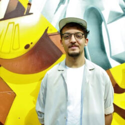 Saib standing in front of graffiti mural outside in a cap and open, grey button-down shirt