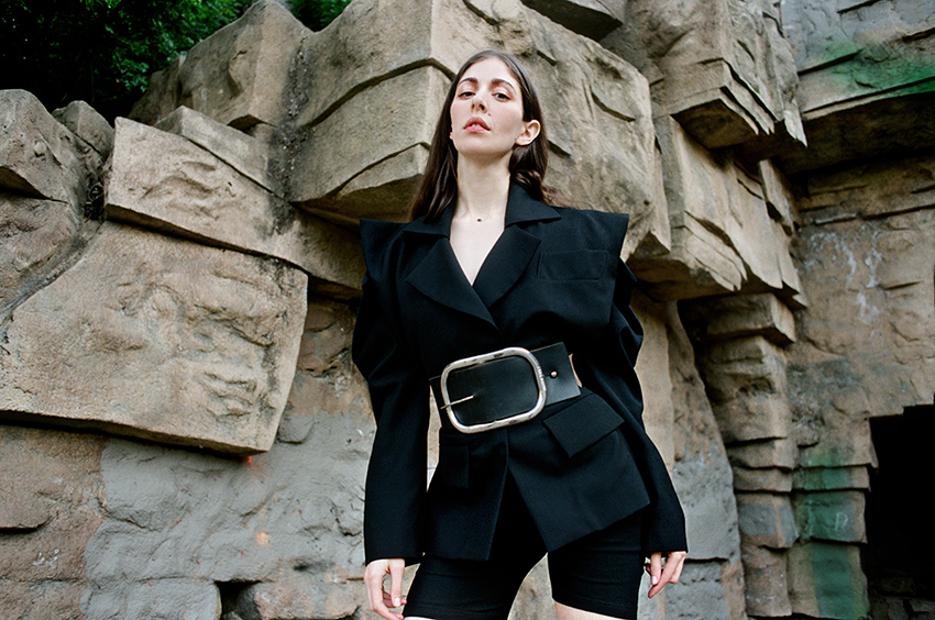 Caroline Polachek dressed in black standing outside by a rock-formed wall