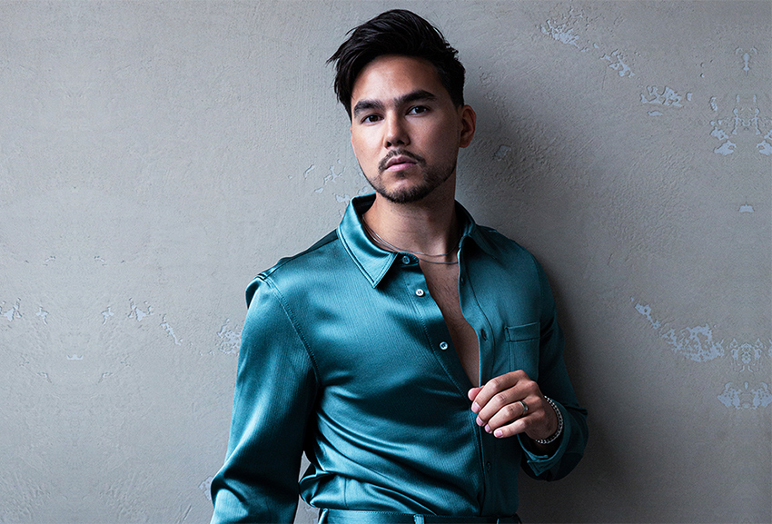 Tyler Shaw standing against a grey wall in a teal silk-like button down shirt.