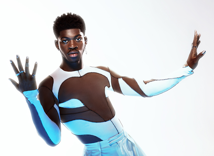 Lil Nas X posing against a bright white background in an black and brown skintight shirt.