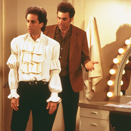 A still from Seinfeld. Jerry's wearing a white blouse with ruffles and puffy sleeves. Kramer stands behind Jerry trying to figure out what he's wearing.