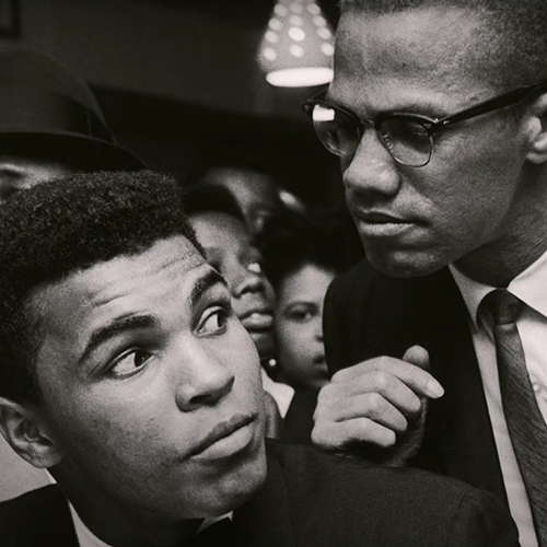 Black and white photo of Malcolm X leaning on Muhammad Ali's shoulders.