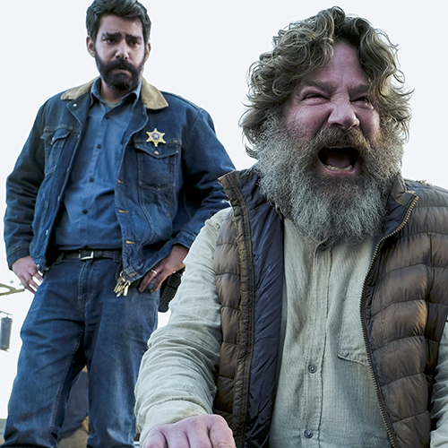 A bearded man yelling with a man dressed in denim with a badge standing behind him.
