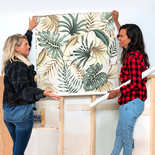 Two women, one blonde, one brunette, holding up a roll of wallpaper against a wall.