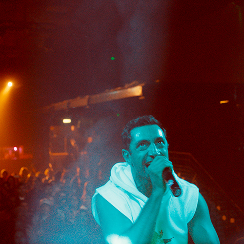 Riz Ahmed in a white shirt holding a microphone.
