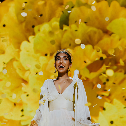 Rupi Kaur in a white dress, standing in front of a giant yellow floral backdrop.