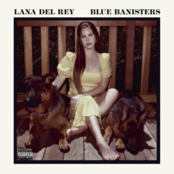 Lana Del Rey's Blue Banisters cover are or Lana sitting on a wood deck, back against a wood rail with german shepherd dogs on either side of her.