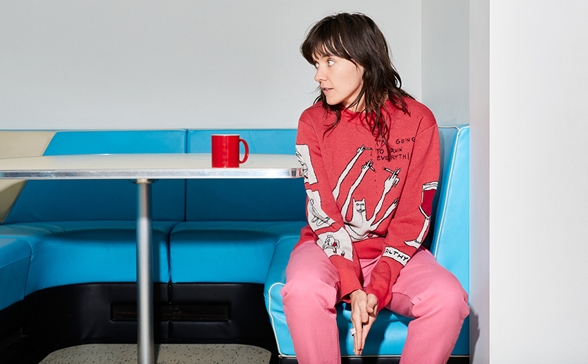 Courtney Barnett dressed in shades of pink and red sitting on the edge of a sky blue bench, looking off to the left.