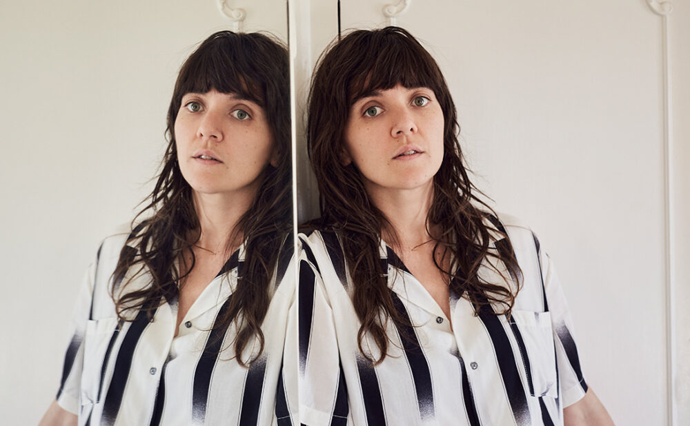 Courtney Barnett leaning against a full length mirror in a black and white top.