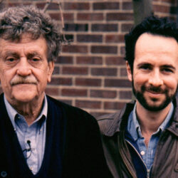 Kurt Vonnegut (left) and Robert Weide (right) standing in front of a brick wall, smiling at the camera.