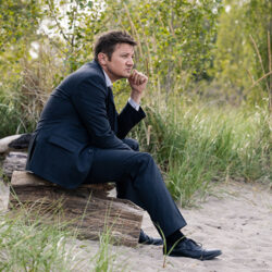 Jeremy Renner in Mayor of Kingstown, sitting on a rock outside in an area with grass and sand.