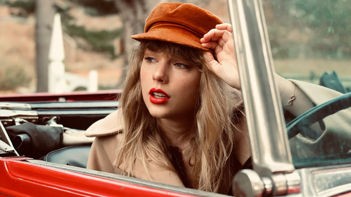 Taylor Swift sitting in a red convertible, top down, wearing a orange-brown hat and camel coloured coat.