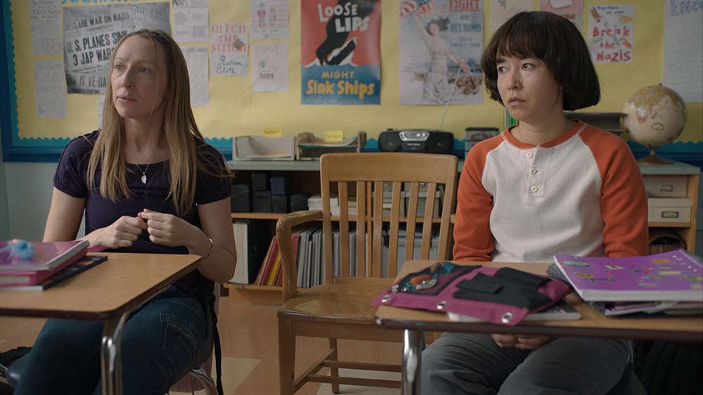Lead actresses from PEN15 sitting in kid's desks